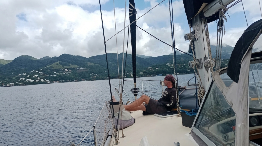 Liveaboard sailor relaxing on a sailboat at anchor in a tropical bay surrounded by green hills.