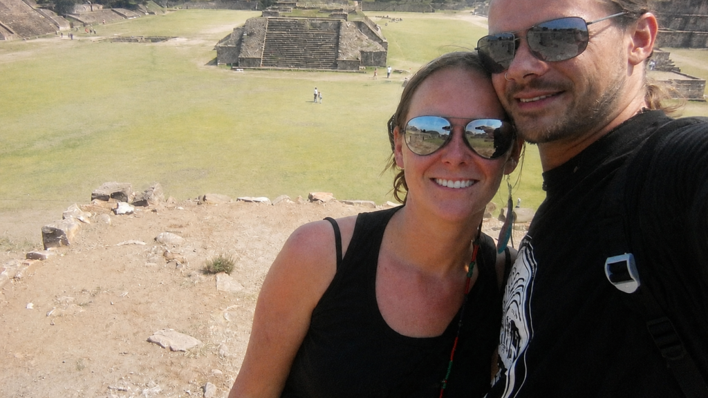 A couple wearing sunglasses pose for a photo at the ancient ruins of Tikal in Guatemala, with a large grassy plaza and stepped stone temples behind them.